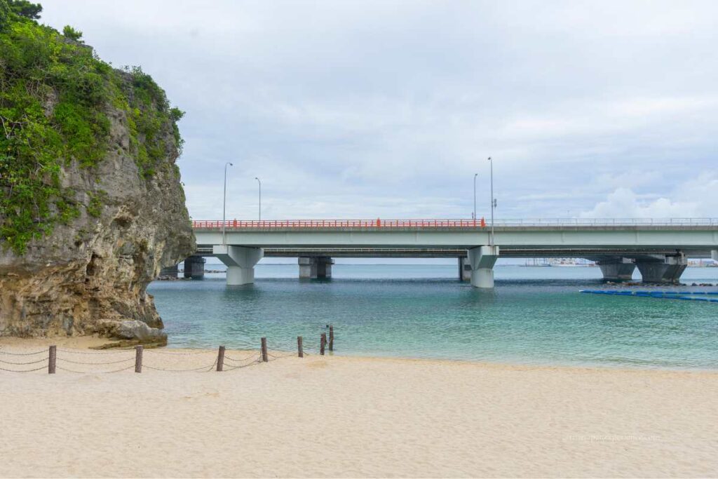 Naminoue Beach in Okinawa with a rocky cliff and the Yui Rail monorail bridge crossing the sea in the background.