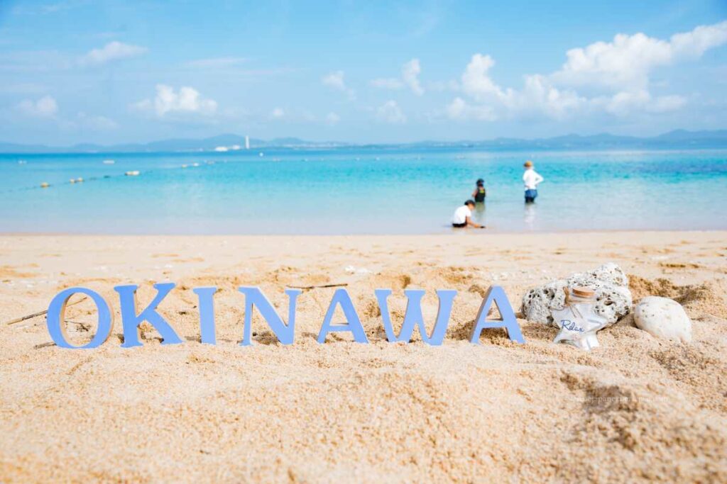 Letters spelling “Okinawa” on a sandy beach with clear blue water and people playing in the sea.