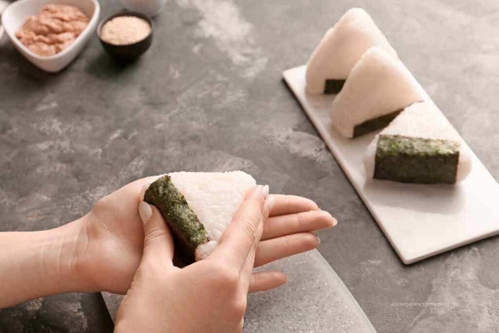 Hands shaping a traditional Japanese onigiri rice ball with seaweed, with more onigiri and ingredients in the background.