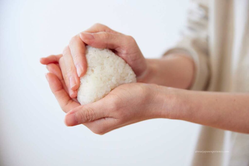 Hands gently shaping a plain white rice ball (onigiri) with a soft background.