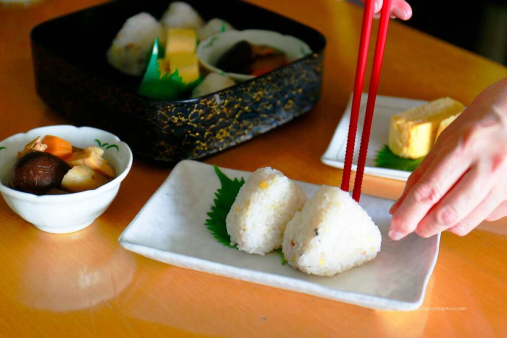 A person placing a rice ball on a plate using red chopsticks, with Japanese side dishes in the background.