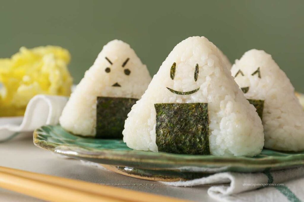Three rice balls (onigiri) with seaweed faces showing different expressions, placed on a green plate.