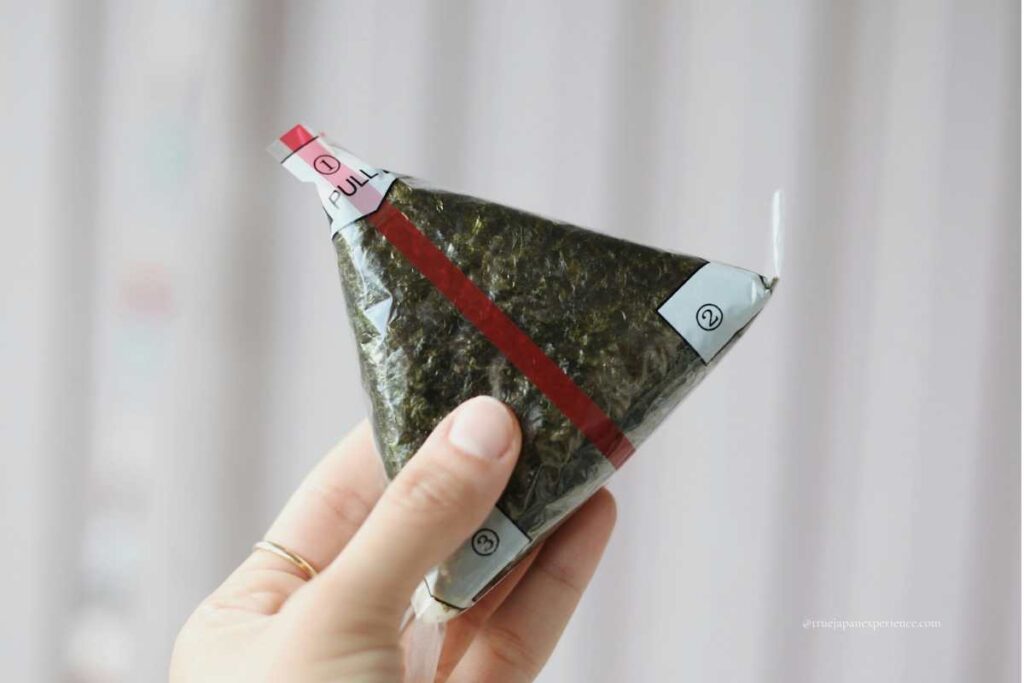 A hand holding a convenience store onigiri wrapped in seaweed with numbered packaging for easy opening.