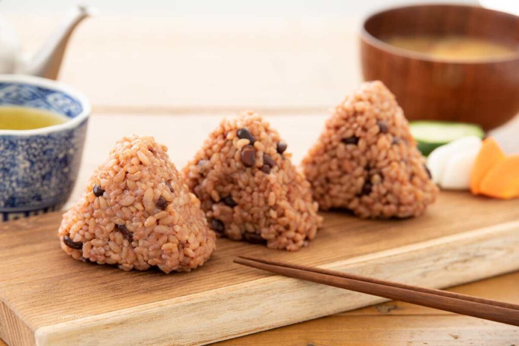 Three red bean rice balls (sekihan onigiri) placed on a wooden tray, with green tea and miso soup in the background.