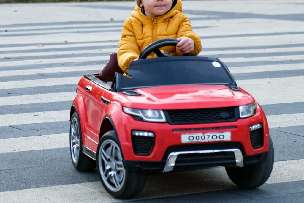 A young child driving a red toy car across a striped pedestrian crossing, wearing a yellow jacket and focused on the steering wheel.