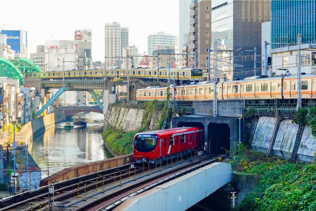 Multiple trains crossing over a canal in central Tokyo, including a red subway train and yellow and orange JR line trains, with city buildings in the background.