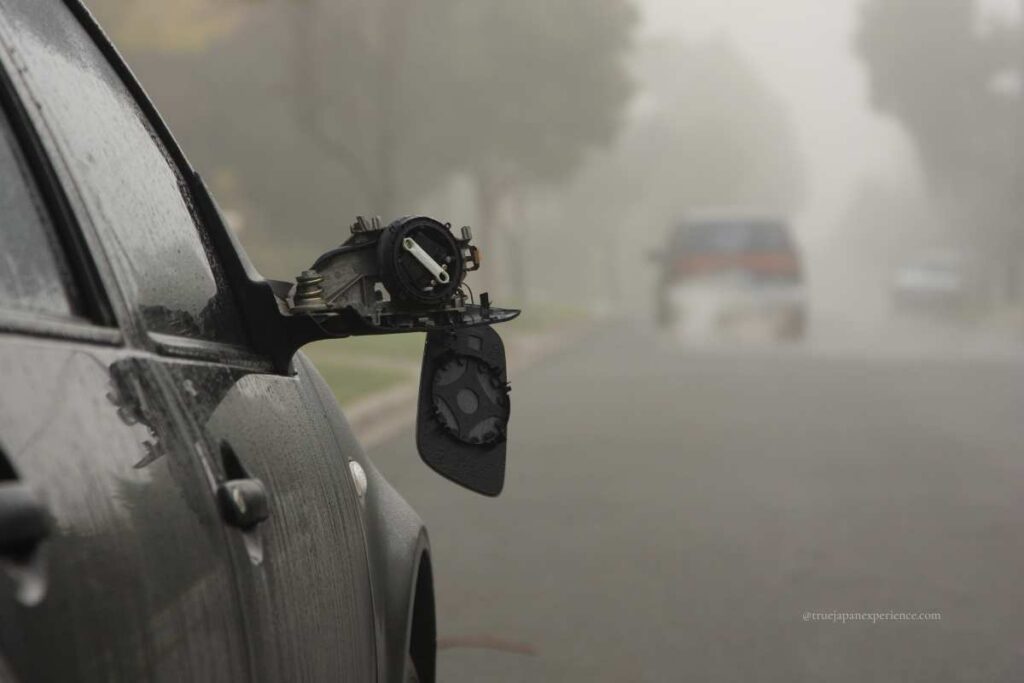 Close-up of a damaged car with a broken side mirror on a foggy road, with other vehicles barely visible in the background.