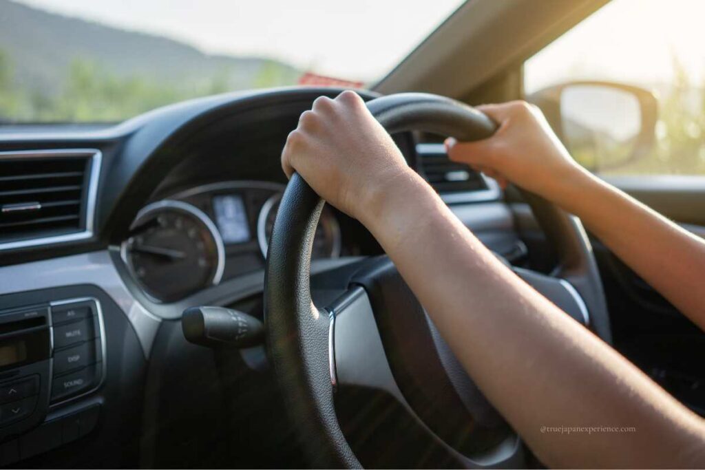 Hands holding a steering wheel in a right-hand drive car, with sunlight streaming through the window—typical of driving in Japan.