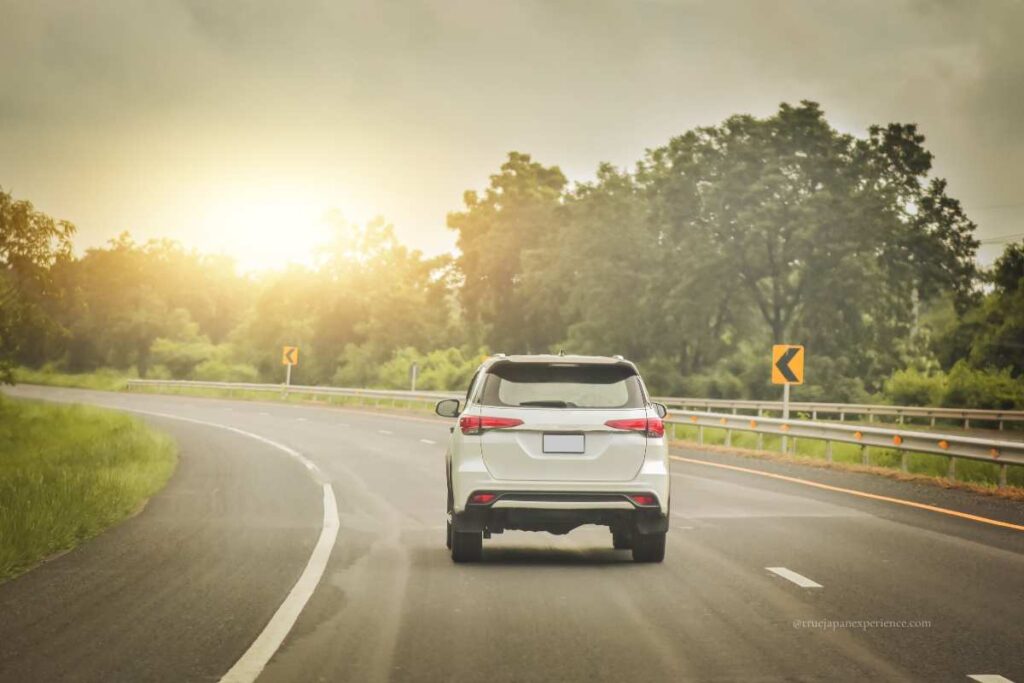 A white SUV driving on the left side of a curving road at sunset in Japan, surrounded by trees and guardrails.