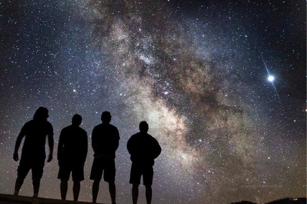 Four people silhouetted against a stunning view of the Milky Way in the night sky, gazing upward in awe.