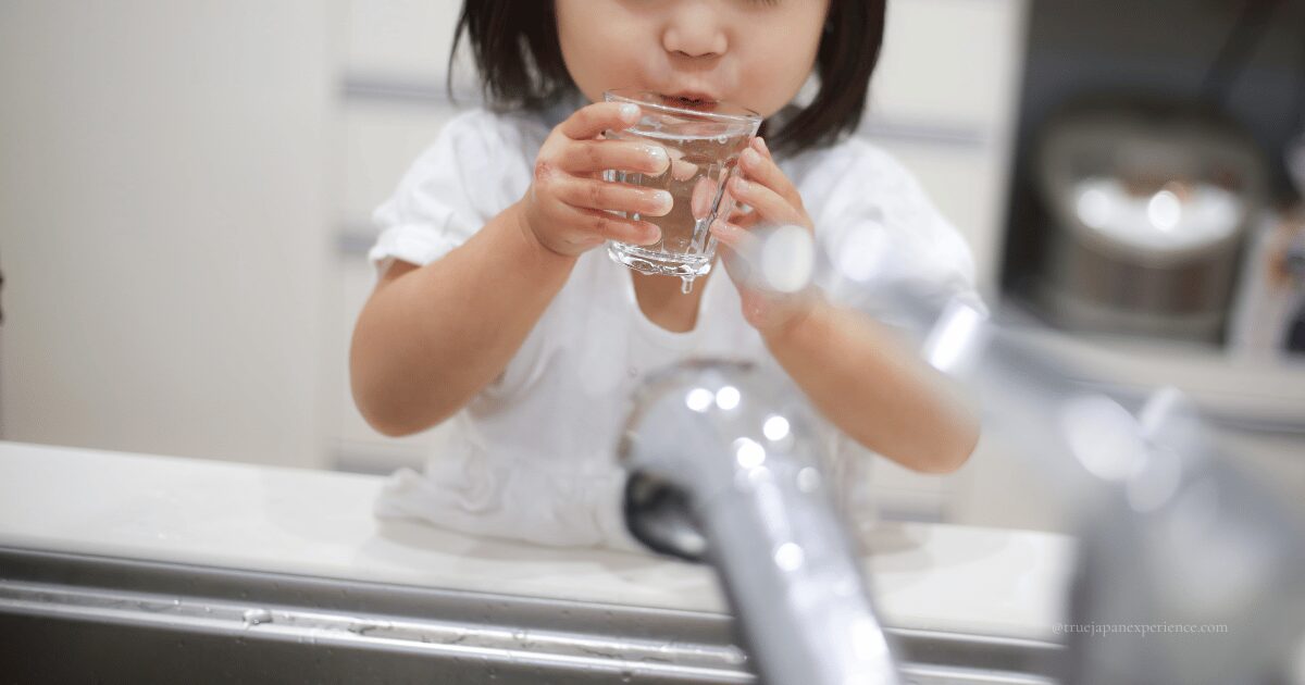 A young child drinking a glass of tap water in a modern Japanese kitchen.