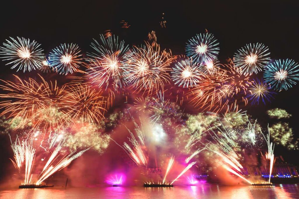A large fireworks display with colorful bursts lighting up the night sky over the water, with reflections and stage lights below.
