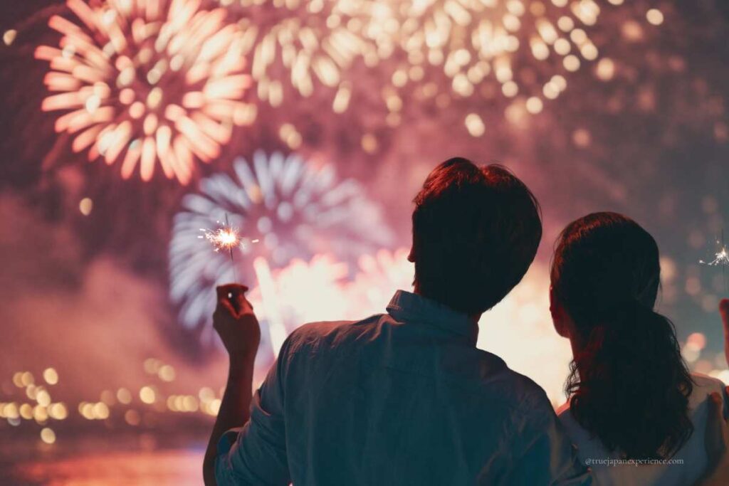 A couple watching fireworks together at night, holding sparklers with colorful fireworks lighting up the sky in front of them.