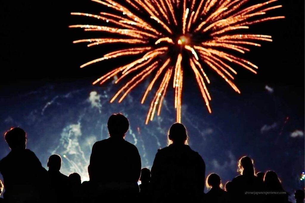 A crowd of people watching a large orange firework burst in the night sky, silhouetted against the glowing light.