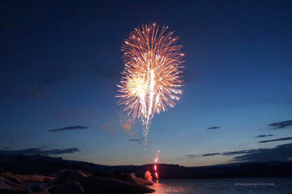 A single firework bursting into the night sky over a calm body of water, with a dark mountain silhouette in the background.