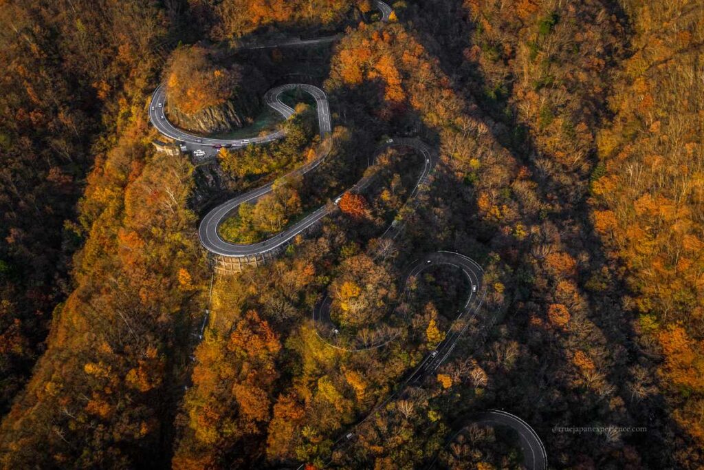 Aerial view of Irohazaka Winding Road in Nikko, surrounded by dense autumn foliage in vibrant shades of red, orange, and yellow.