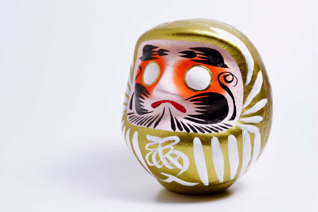 Close-up of a golden Daruma doll with bold black and red facial features on a plain white background.