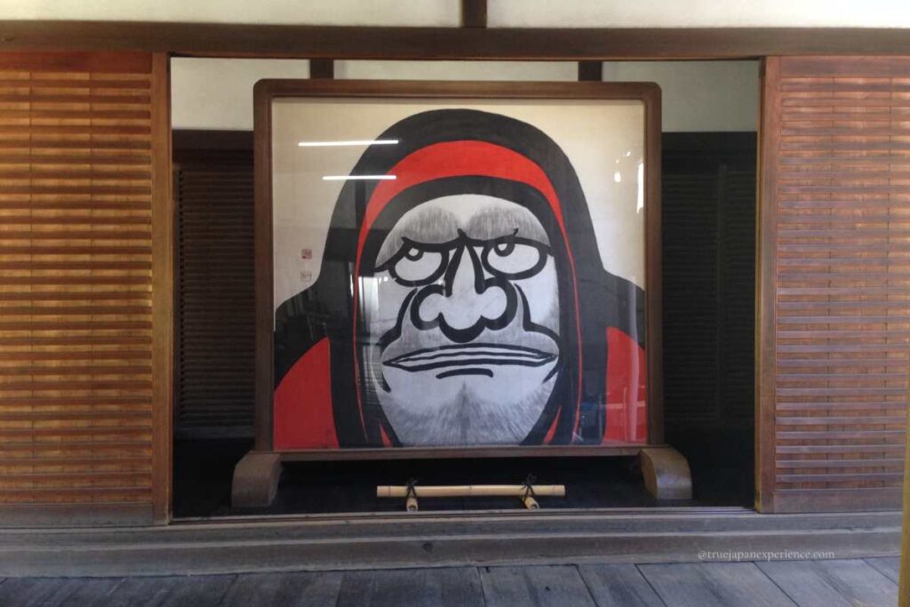 Large painting of Bodhidharma (Daruma) with a red hood displayed inside a traditional Japanese temple hall.