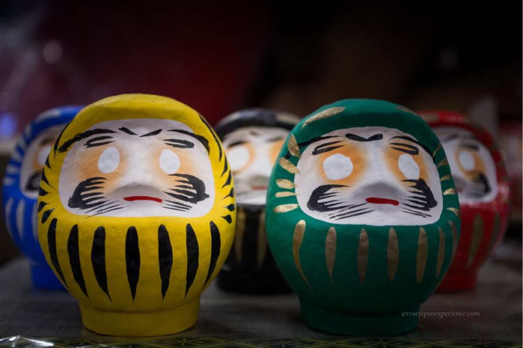 Close-up of yellow, green, blue, and red Daruma dolls displayed side by side on a table.
