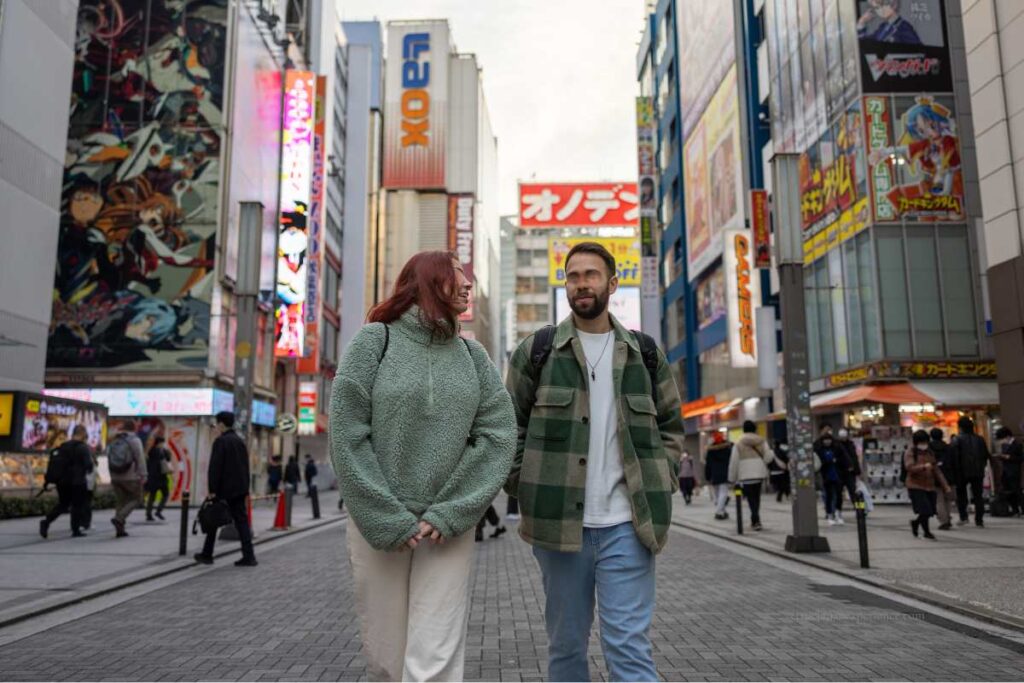 Two foreign tourists walking through a busy shopping street in Akihabara, Tokyo, surrounded by colorful anime billboards and electronics stores.