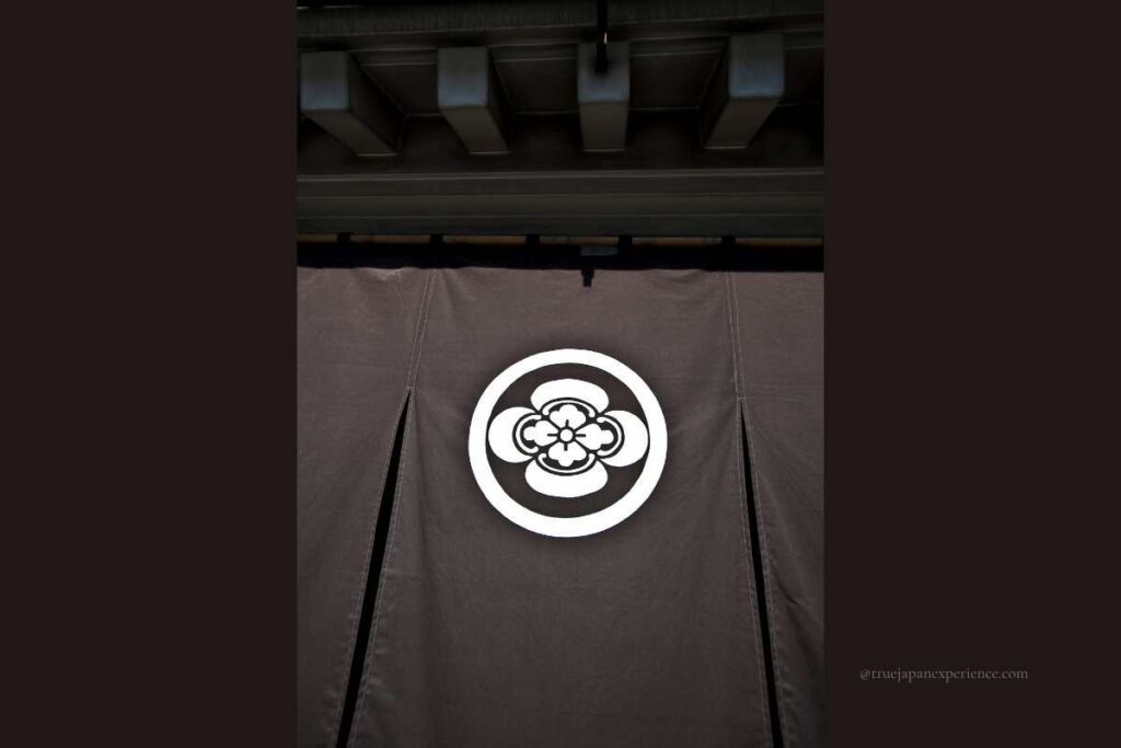 Japanese family crest (kamon) featuring a stylized flower inside a circle, displayed on a dark fabric curtain (noren) hanging at the entrance of a traditional building.