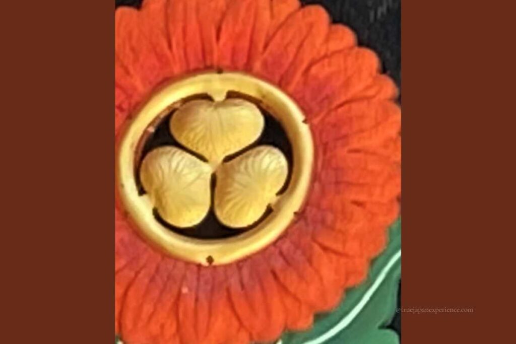Decorative fitting on a Shinto shrine gate featuring a gold family crest (kamon) of three hollyhock leaves inside a circle, set against a red floral background.