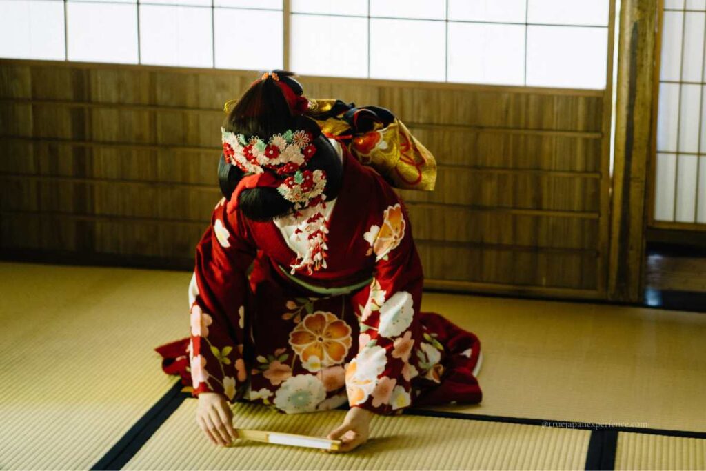 Japanese woman in a red kimono bowing on tatami floor, holding a folded sensu fan in a traditional setting