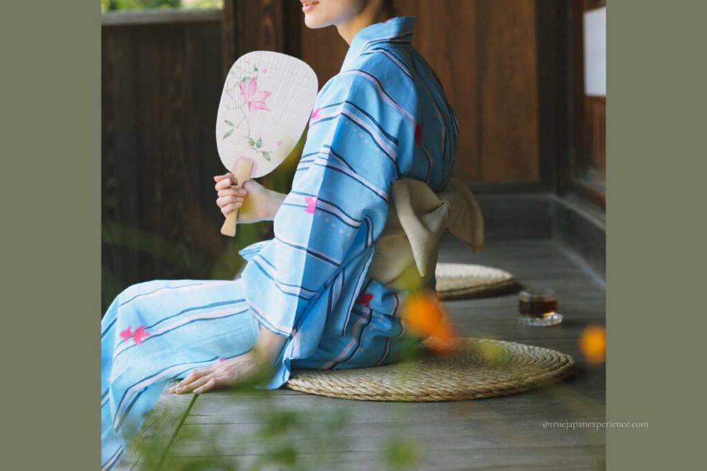 Japanese woman in a blue yukata sitting on a porch, holding a traditional uchiwa hand fan with floral design