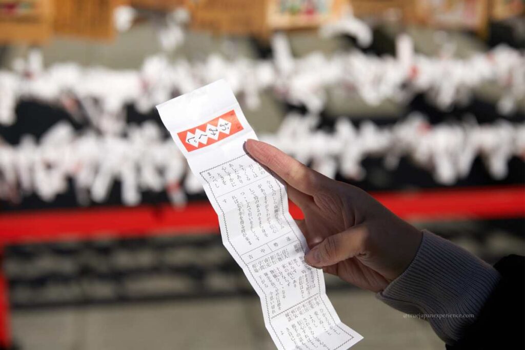 A close-up of a person holding a Japanese omikuji paper at a shrine or temple, with omikuji tied in the background.