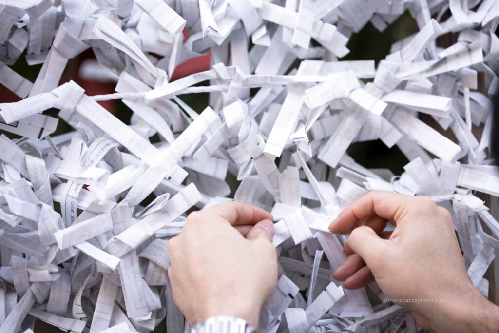 Hands tying an omikuji paper to a designated rack at a Japanese shrine.