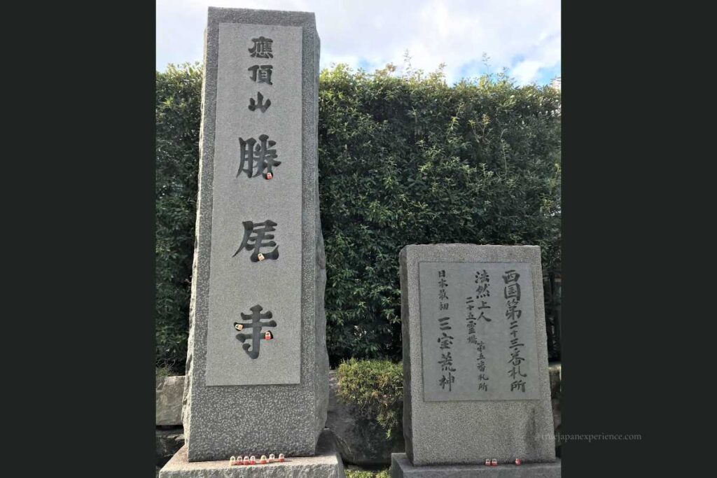 Stone monument at the entrance of Katsuoji Temple with large carved kanji, decorated with small daruma dolls placed in the grooves.