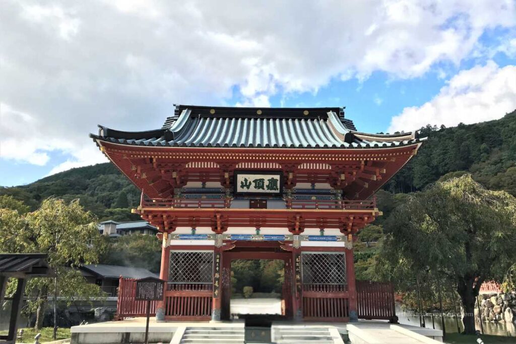 Front view of the grand entrance gate at Katsuoji Temple in Osaka, with red pillars, a blue-tiled roof, and forested hills behind.