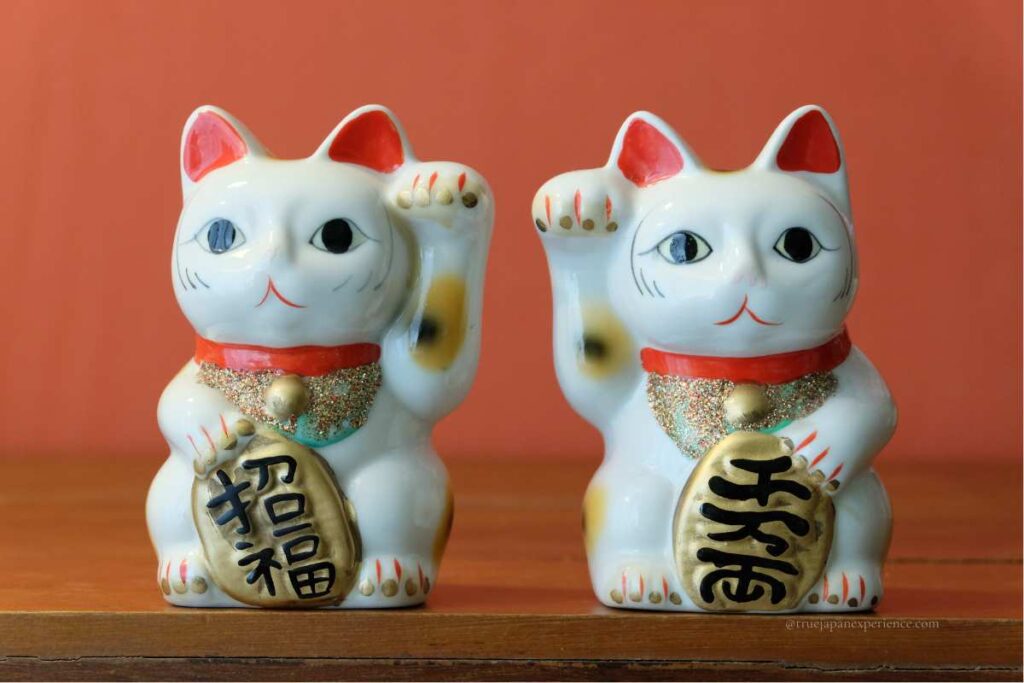 Two traditional white Maneki-neko statues with raised paws, wearing red collars and holding gold koban coins with Japanese characters.