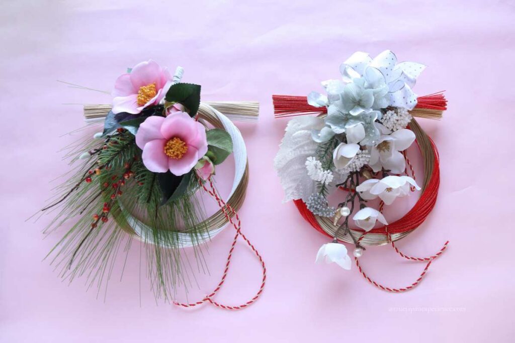 Two decorative Japanese mizuhiki wreaths on a pink background, one with pink camellia flowers and pine, the other with white flowers and red cords.