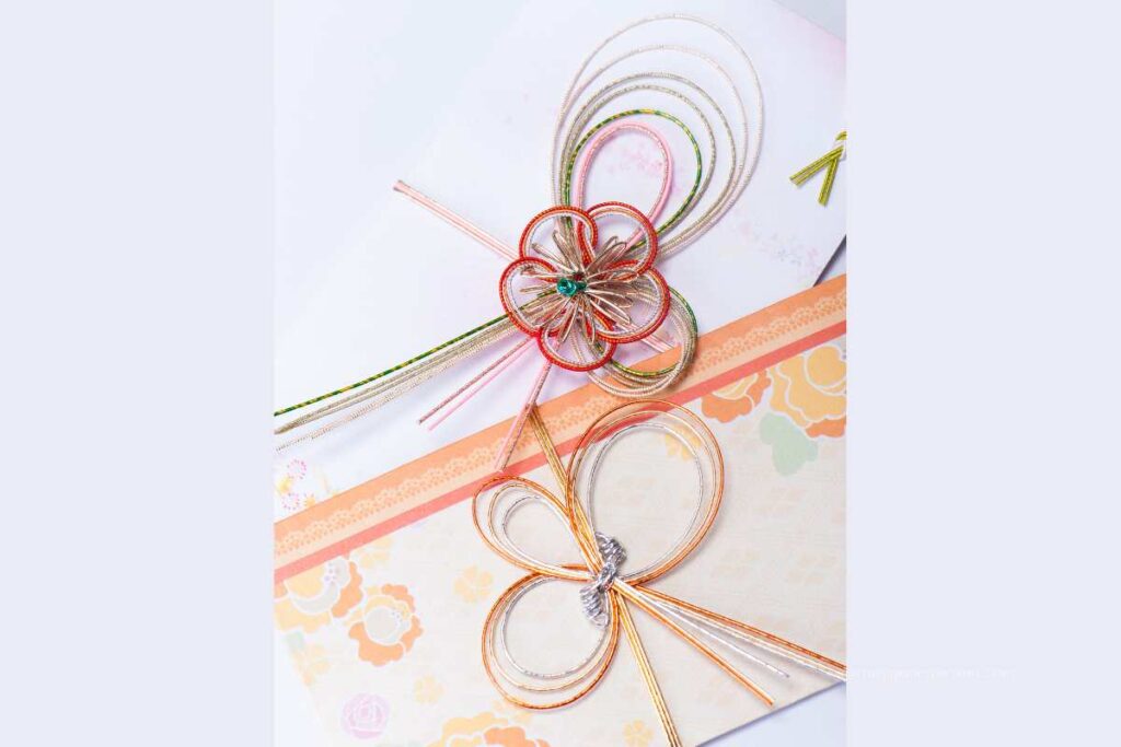 Two Japanese envelopes decorated with colorful mizuhiki cords, one shaped like a flower and the other tied in a bow, placed on patterned paper.
