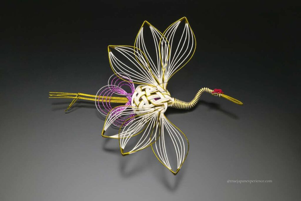 A traditional Japanese mizuhiki decoration shaped like a crane, made with gold, white, and purple cords, placed on a dark background.