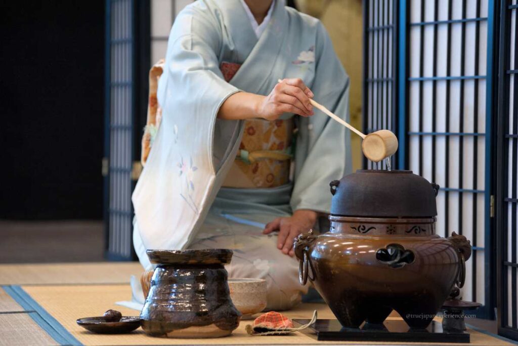 A person in a light blue kimono performing a traditional Japanese tea ceremony, carefully pouring hot water with a bamboo ladle.