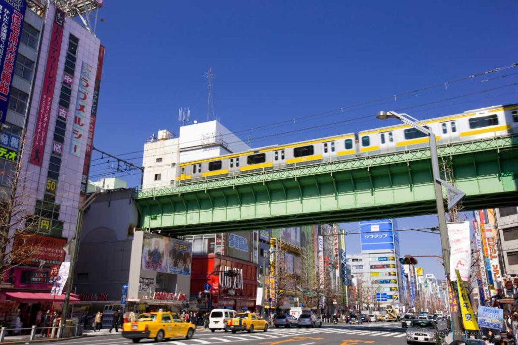 A yellow JR train crossing a green bridge above the busy streets of Akihabara, Tokyo.