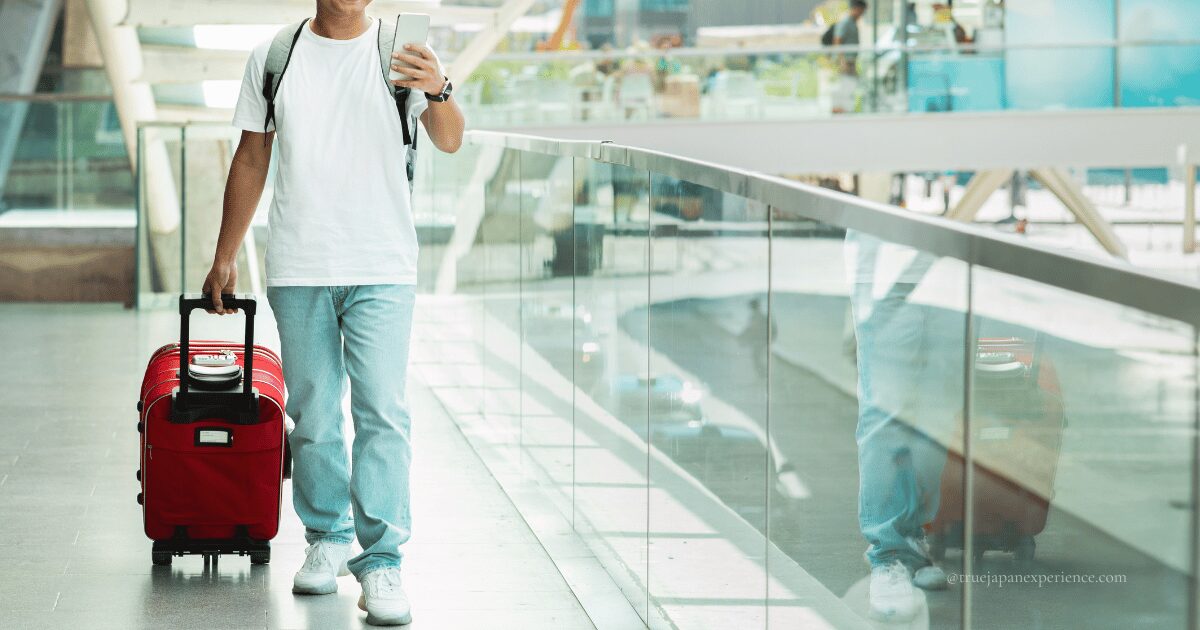 Traveler in casual clothes pulling a red suitcase while checking a smartphone at an airport.