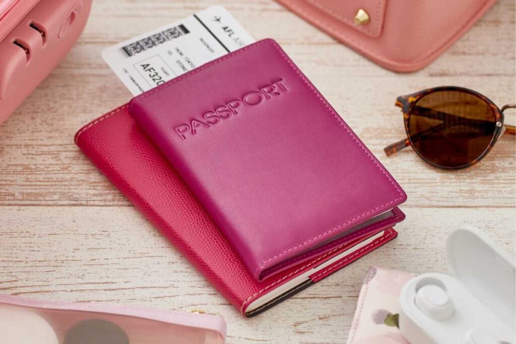 A pink passport holder with a boarding pass on a wooden table, next to sunglasses and a suitcase
