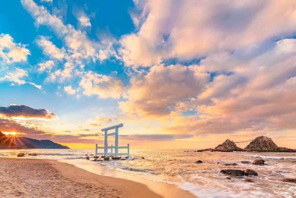 A white torii gate standing in the sea at sunset, with the Married Couple Rocks (Meoto Iwa) in the background in Japan.
