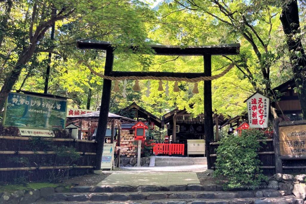 A black wooden torii gate with a sacred rope in front of Nonomiya Shrine, surrounded by green trees in Kyoto.