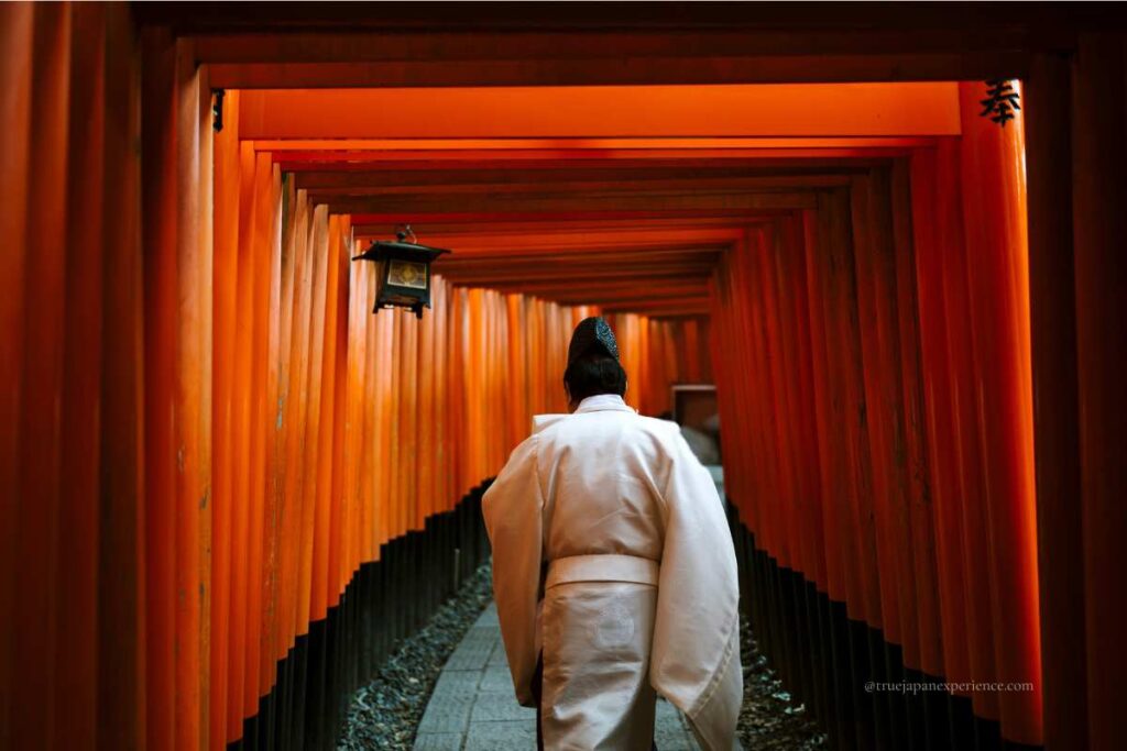 A Shinto priest in white robes walking through a tunnel of red torii gates at Fushimi Inari Shrine in Kyoto.