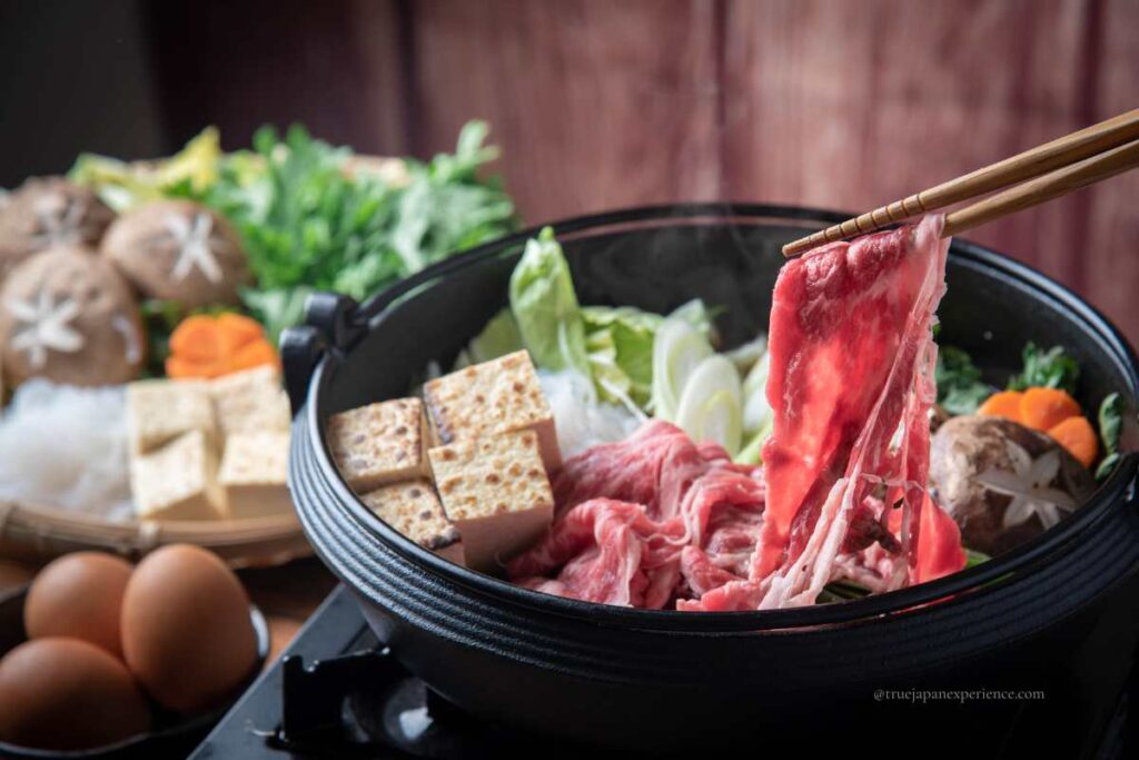A Japanese sukiyaki hot pot with vegetables, tofu, and thin slices of beef being cooked and dipped into raw egg before eating